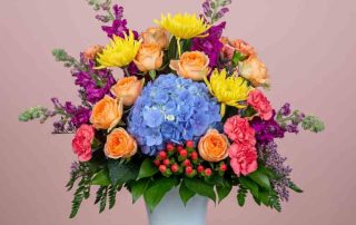 Send joy with this bright and festive arrangement featuring bright blue hydrangea, hot pink spray roses, peach carnations, giant yellow mums, statice, hypericum and multiple types of accenting greens.